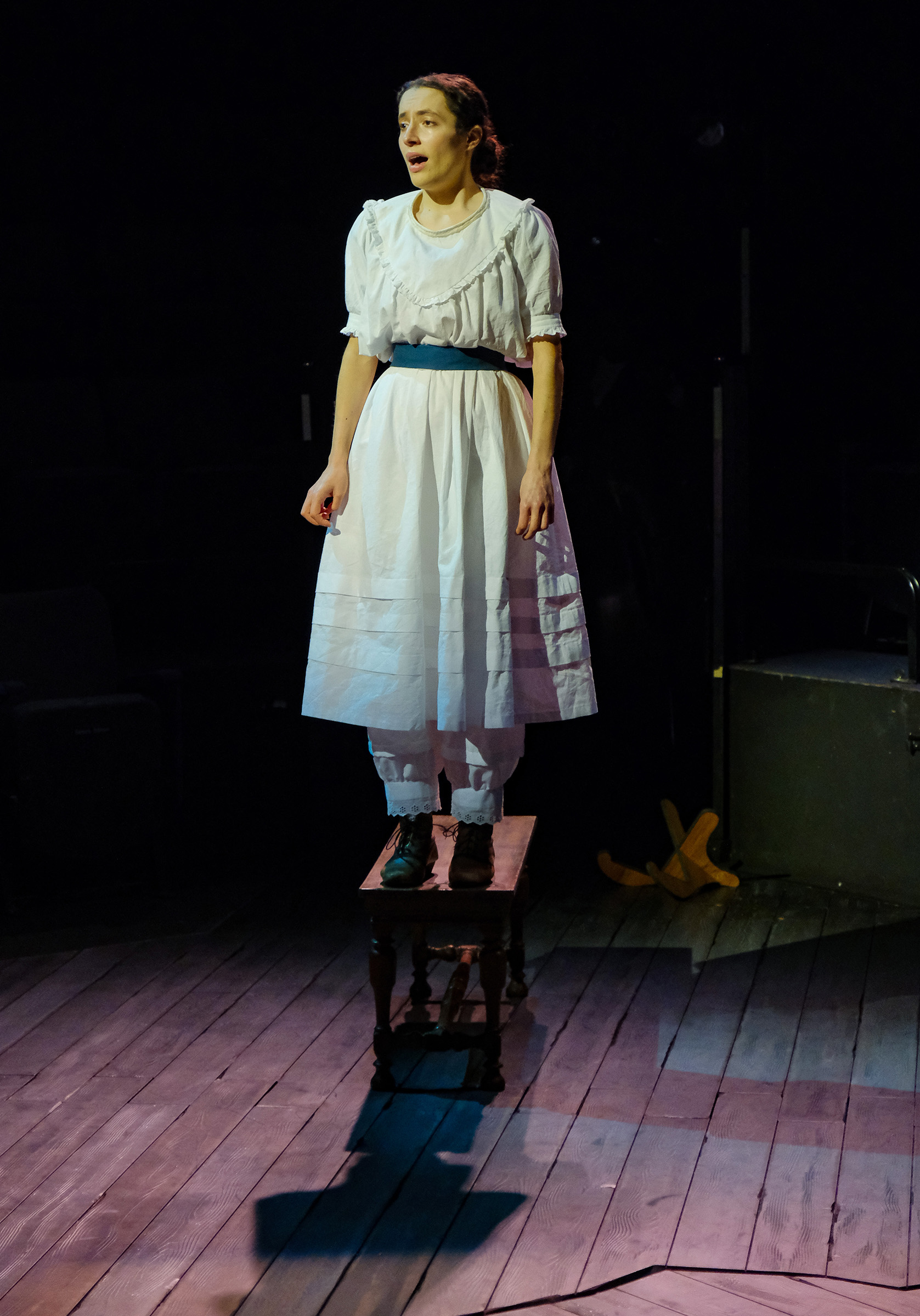 theatre performance image: Jane Eyre in a white dress is standing alone on a bench