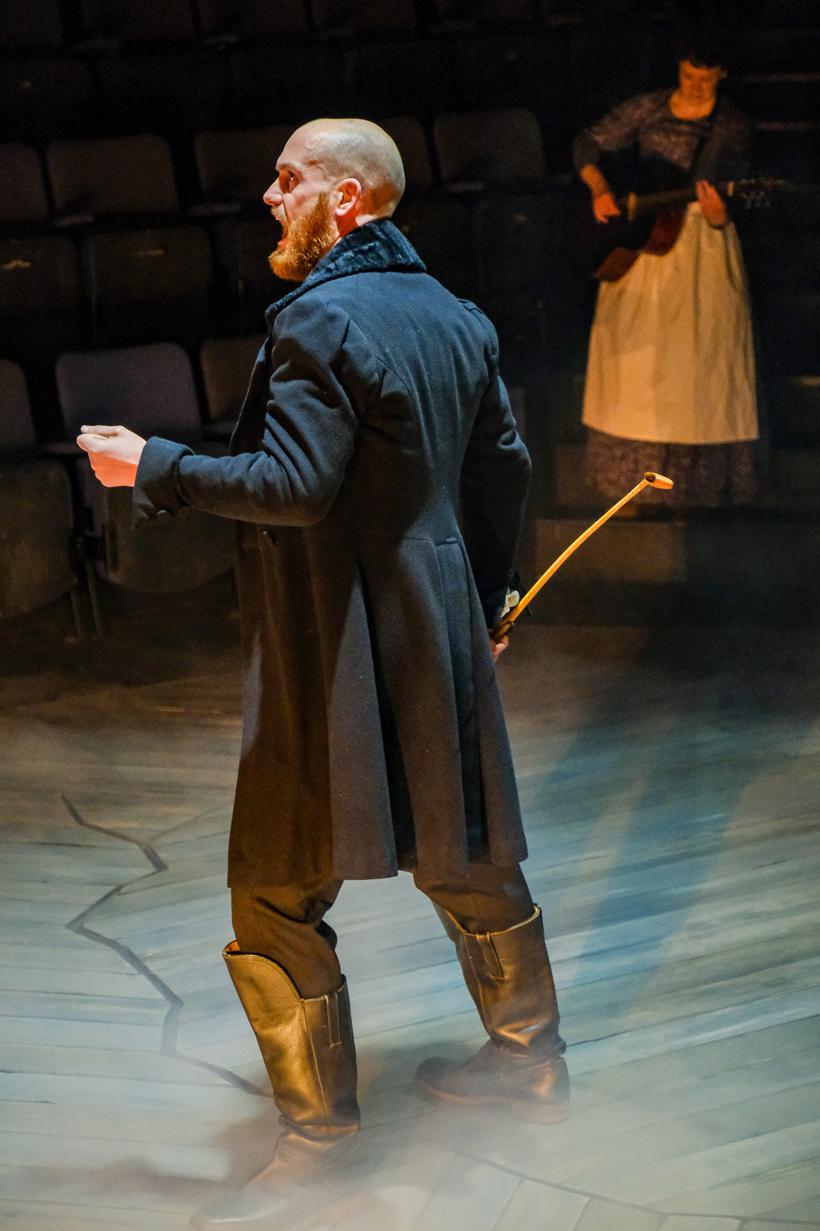 theatre performance image: Rochester stands centre stage dressed all in black. There is another performer playing guitar in the background.